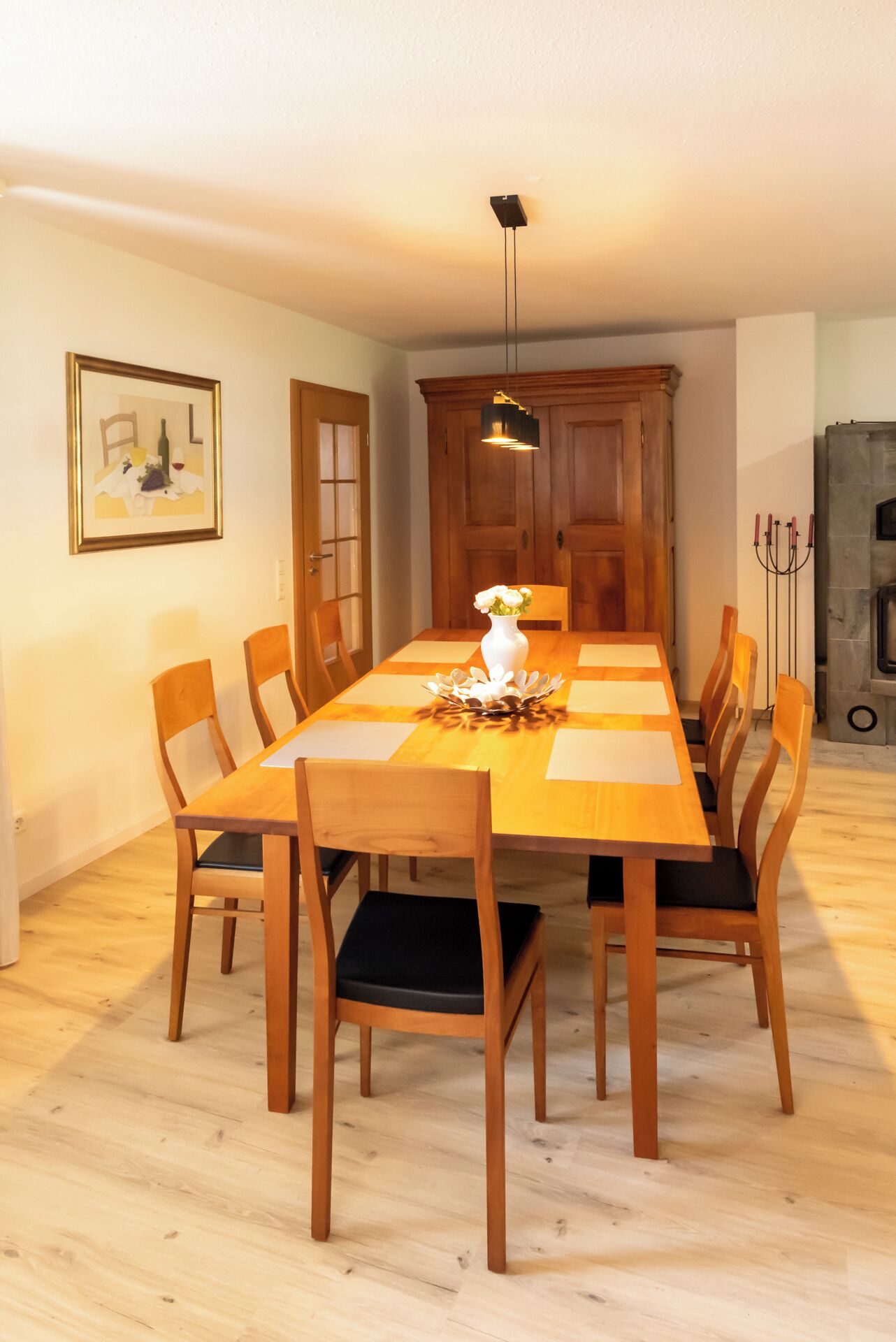 A dining room with a long wooden table, six chairs, a vase with flowers, and a painting on the wall.