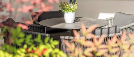 Un área de estar al aire libre con una mesa redonda y sillas, una planta en maceta y una ventana al fondo.