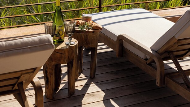 Eine Holzterrasse mit zwei Liegestühlen und einem kleinen Tisch, mit Blick auf eine Küstenlandschaft mit viel Grün und dem Meer.