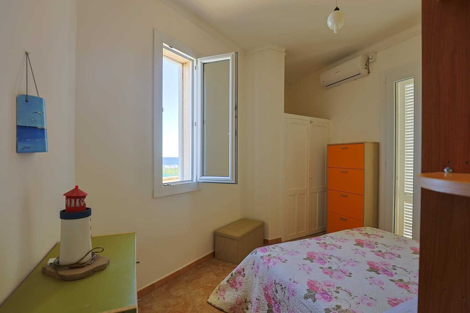 Une chambre à coucher avec un lit, une fenêtre, une armoire, une petite table sur laquelle est posé un modèle de phare, et un radiateur.