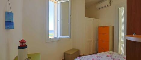 Une chambre à coucher avec un lit, une fenêtre, une armoire, une petite table sur laquelle est posé un modèle de phare, et un radiateur.