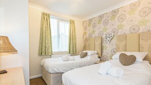Quarto de hotel com duas camas, janela com cortinas, luminária e papel de parede floral.