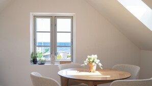 Ein Essbereich mit einem runden Tisch, vier Stühlen, einem Fenster mit Meerblick und einem Blumentopf auf der Fensterbank.