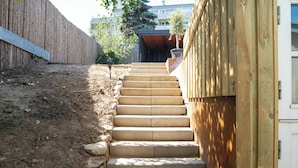 Eine Betontreppe führt zu einem Hauseingang, umgeben von einem Holzzaun und einem Garten mit einem Blumentopf.