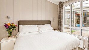 Une chambre à coucher avec un lit, une table de chevet, un vase de fleurs, une fenêtre avec des rideaux et une vue sur l’extérieur.
