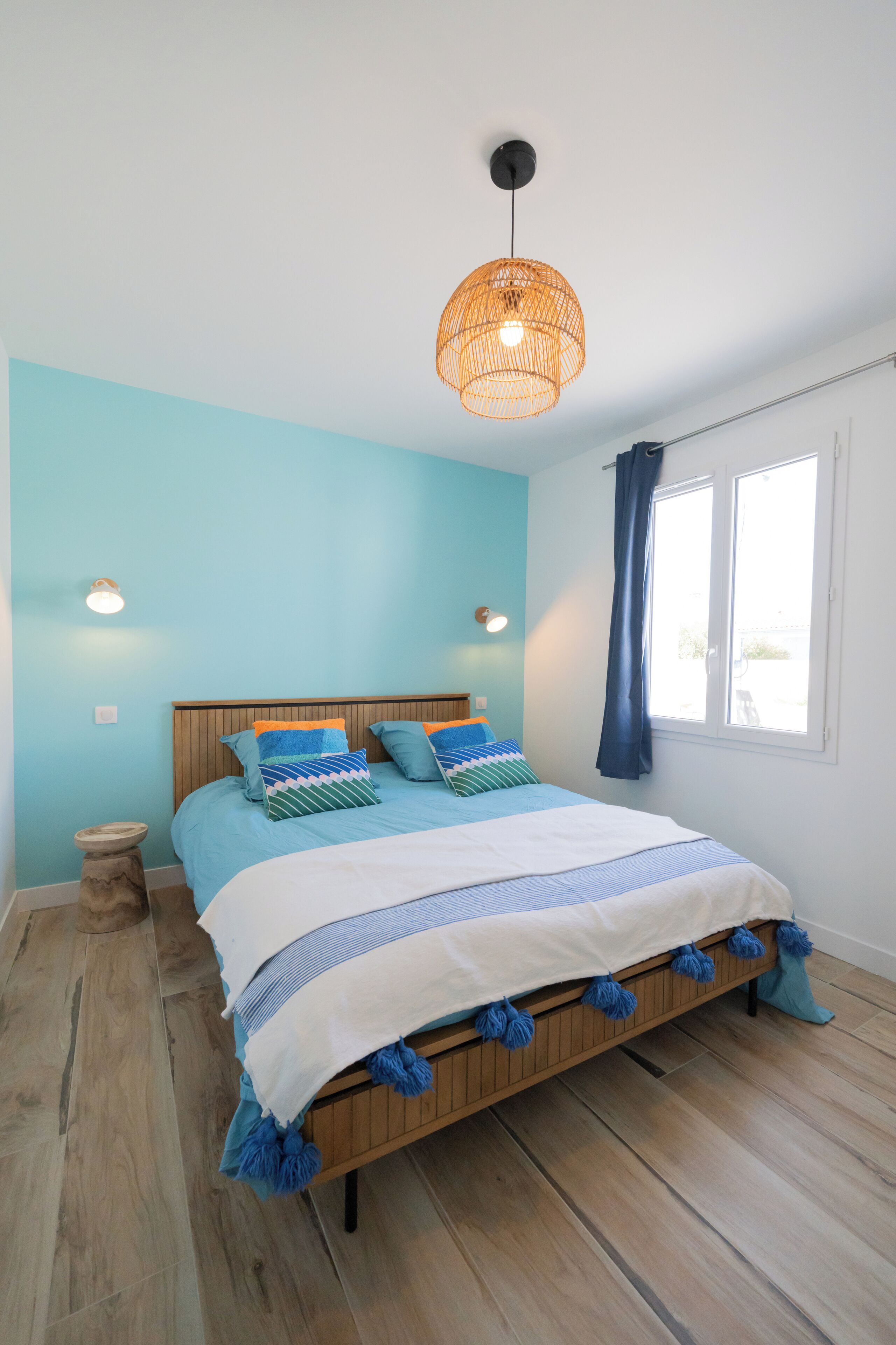 A bedroom with a wooden bed, blue bedding, a hanging rattan light fixture, and a window with curtains.