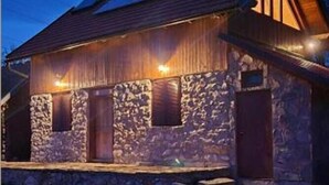 Un bâtiment rustique en pierre, doté d’un toit en pente et de poutres en bois, avec de nombreuses portes et fenêtres, éclairé par un système d’éclairage extérieur.