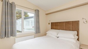A bedroom with a bed, curtains, a window, a radiator, a bedside table, and a wall-mounted light fixture. - 2 bedroom accommodation in Talacre, Holywell (Talacre, Holywell)