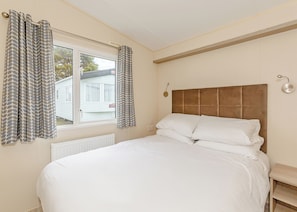 A bedroom with a bed, curtains, a window, a radiator, a bedside table, and a wall-mounted light fixture. - 2 bedroom accommodation in Talacre, Holywell (Talacre, Holywell)