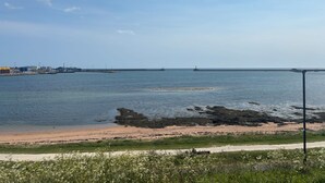 A coastal view with a sandy beach, calm water, and industrial structures in the distance. - Beachview  (Peterhead)