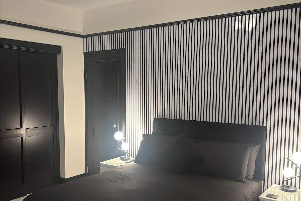 A modern bedroom with a large bed, a nightstand with a lamp, a wardrobe, and a ceiling fan.