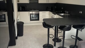 A modern kitchen with white cabinets, a black countertop, a built-in oven, a microwave, and a coffee maker. There are three black bar stools and a grey sofa. - Beachview  (Peterhead)