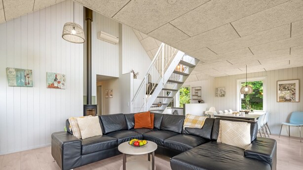 Ein geräumiges Wohnzimmer mit einem schwarzen Ledersofa, einem runden Couchtisch mit Früchten, einer Treppe und einem Essbereich mit weißem Tisch und Stühlen.