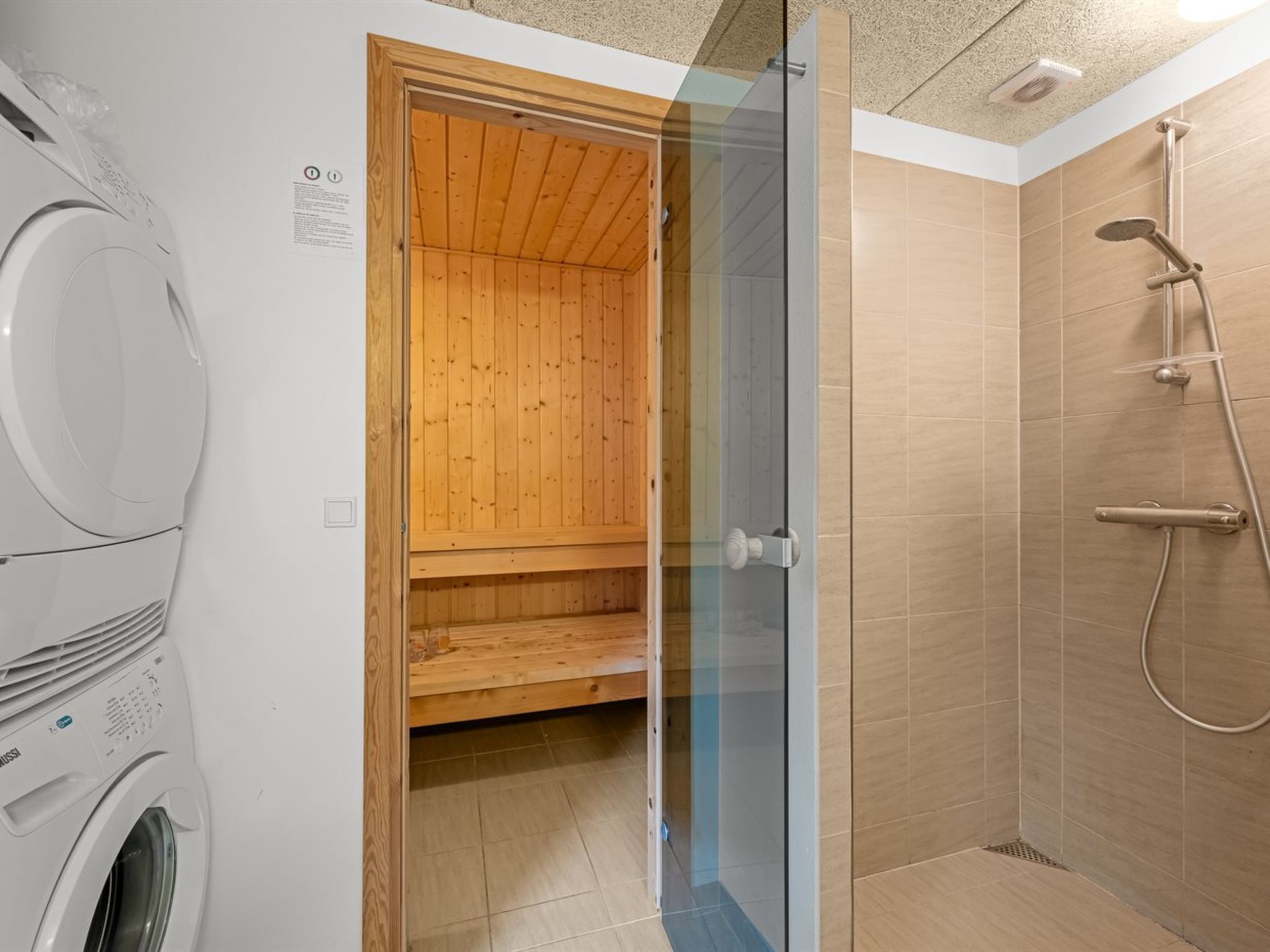 Een badkamer met een douche in glazen cabine, een houten sauna en een wasmachine.