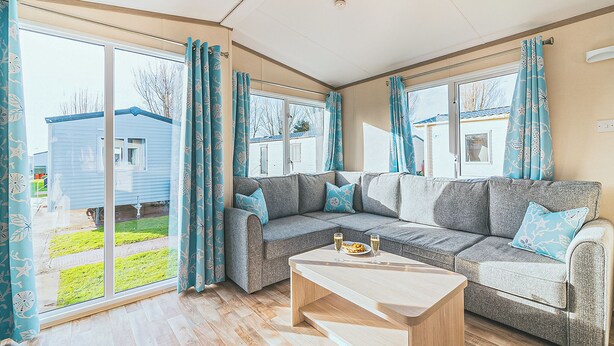 A spacious living room with a large window, a sofa set, and a wooden coffee table. - 3 bedroom accommodation in Talacre, Holywell (Talacre, Holywell)