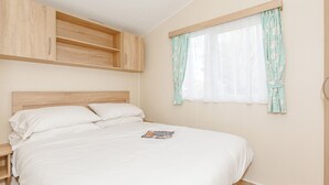 A bedroom with a wooden bed frame, white bedding, a nightstand, a window with curtains, and built-in wooden cabinets above the bed. - 3 bedroom accommodation in Talacre, Holywell (Talacre, Holywell)