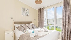 Un dormitorio con cama, mesita de noche, reloj de pared, ventana con cortinas y vista a una casa.