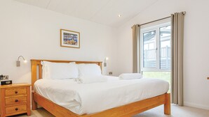 A bedroom with a wooden bed, a nightstand, a wall-mounted lamp, and a window with curtains. - Fresh and contemporary styled lodge. (Axminster)