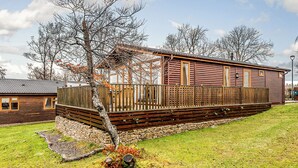 Eine Blockhütte mit einer Holzterrasse, einem Steinfundament und einem Baum im Vordergrund.