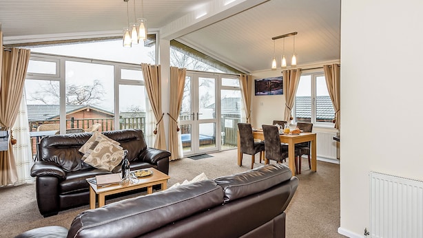 Ein geräumiges Wohnzimmer mit Ledersofa, Esstisch und Stühlen, großen Fenstern mit Vorhängen sowie einem Fernseher an der Wand.