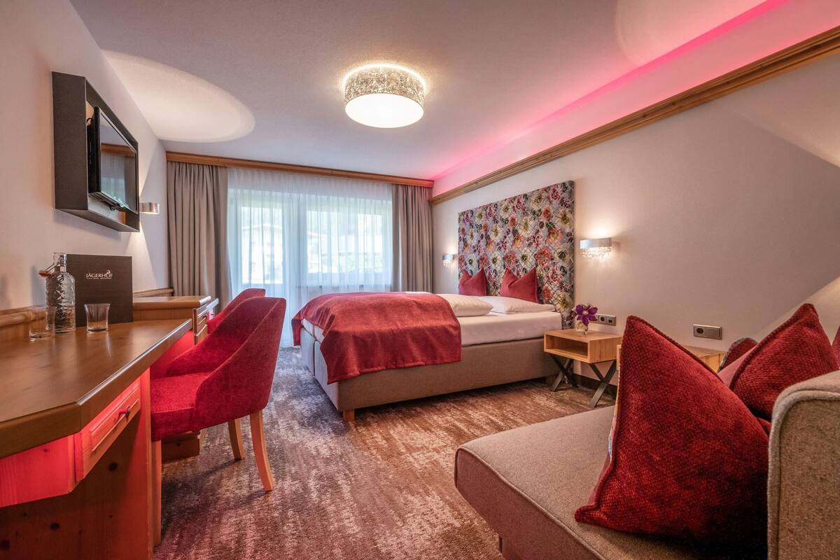 A hotel room with a bed, a desk with a chair, a sofa, a large window with curtains, a wall-mounted TV, and a decorative wall piece.
