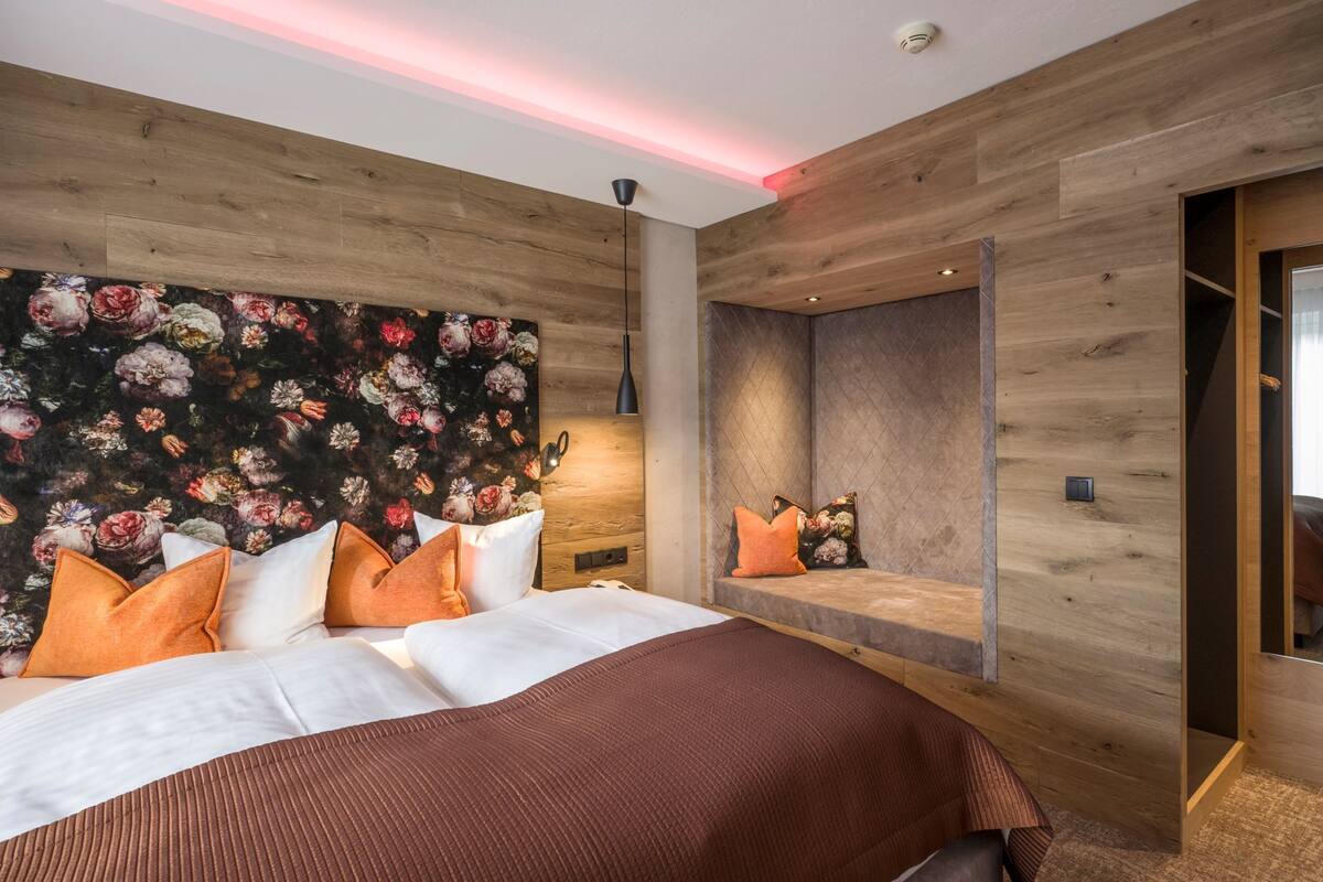 A modern bedroom with a large bed, a floral wallpaper headboard, a bench with cushions, and a wooden wall design.