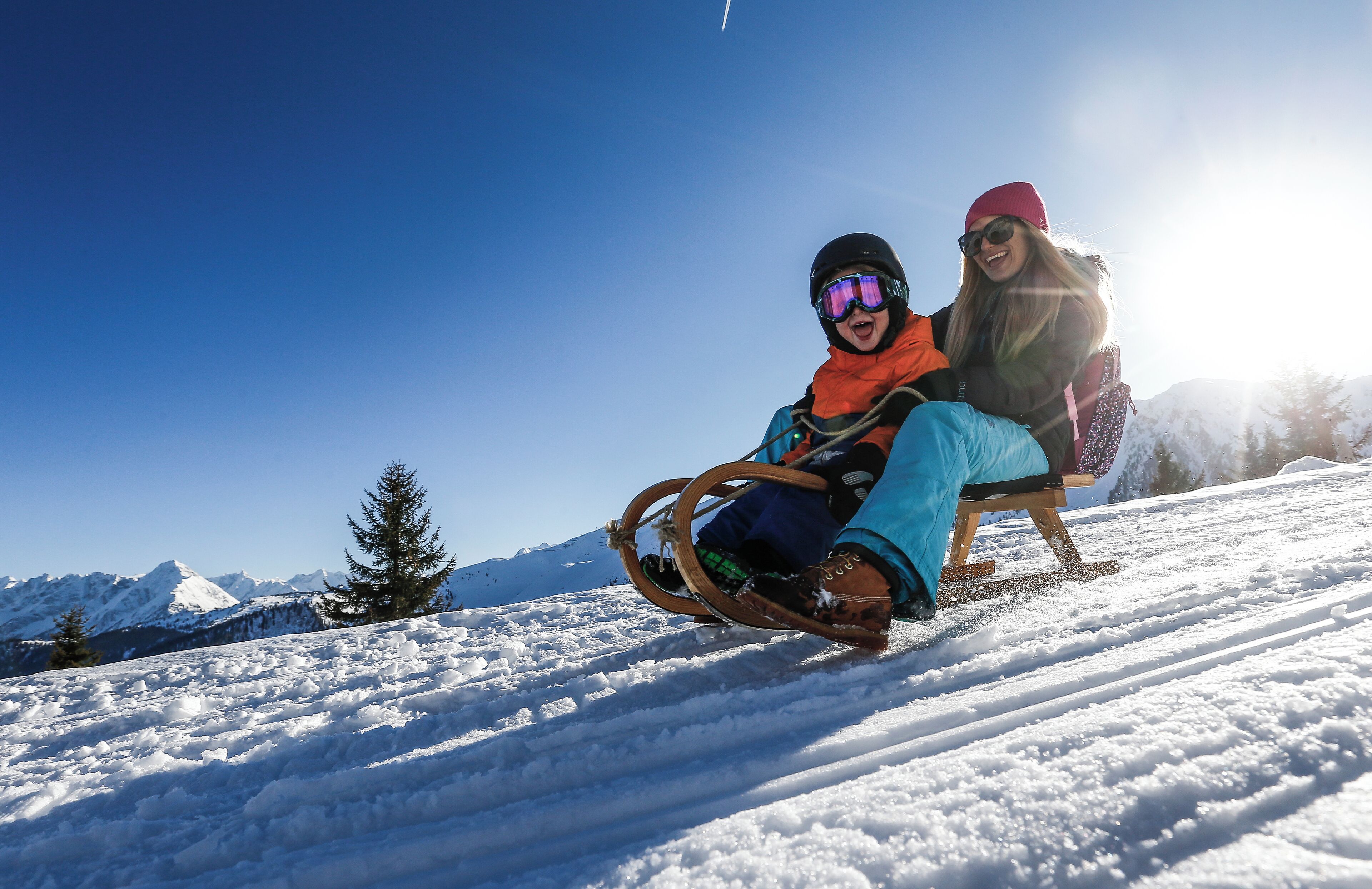 Two people sledding down a snowy slope with a clear sky and mountains in the background.