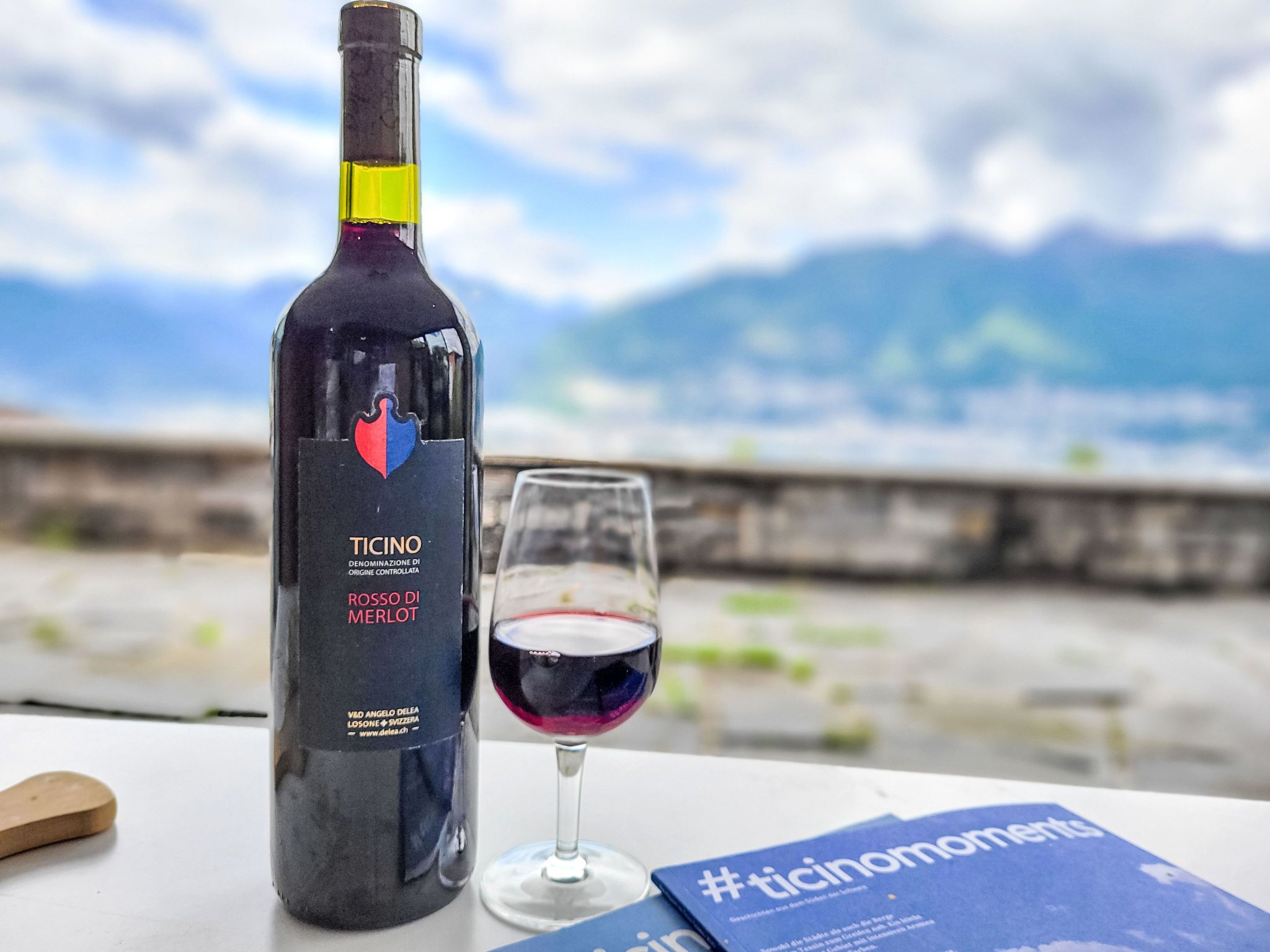 Una bottiglia di vino rosso Merlot Ticinomoments e un bicchiere di vino su un tavolo con vista panoramica sulle montagne.