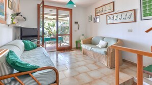 A room with a sofa, a wicker chair, a television, and a table. There is a door leading to an outdoor area with plants. - Sunny House by Interhome (Marina di Campo)