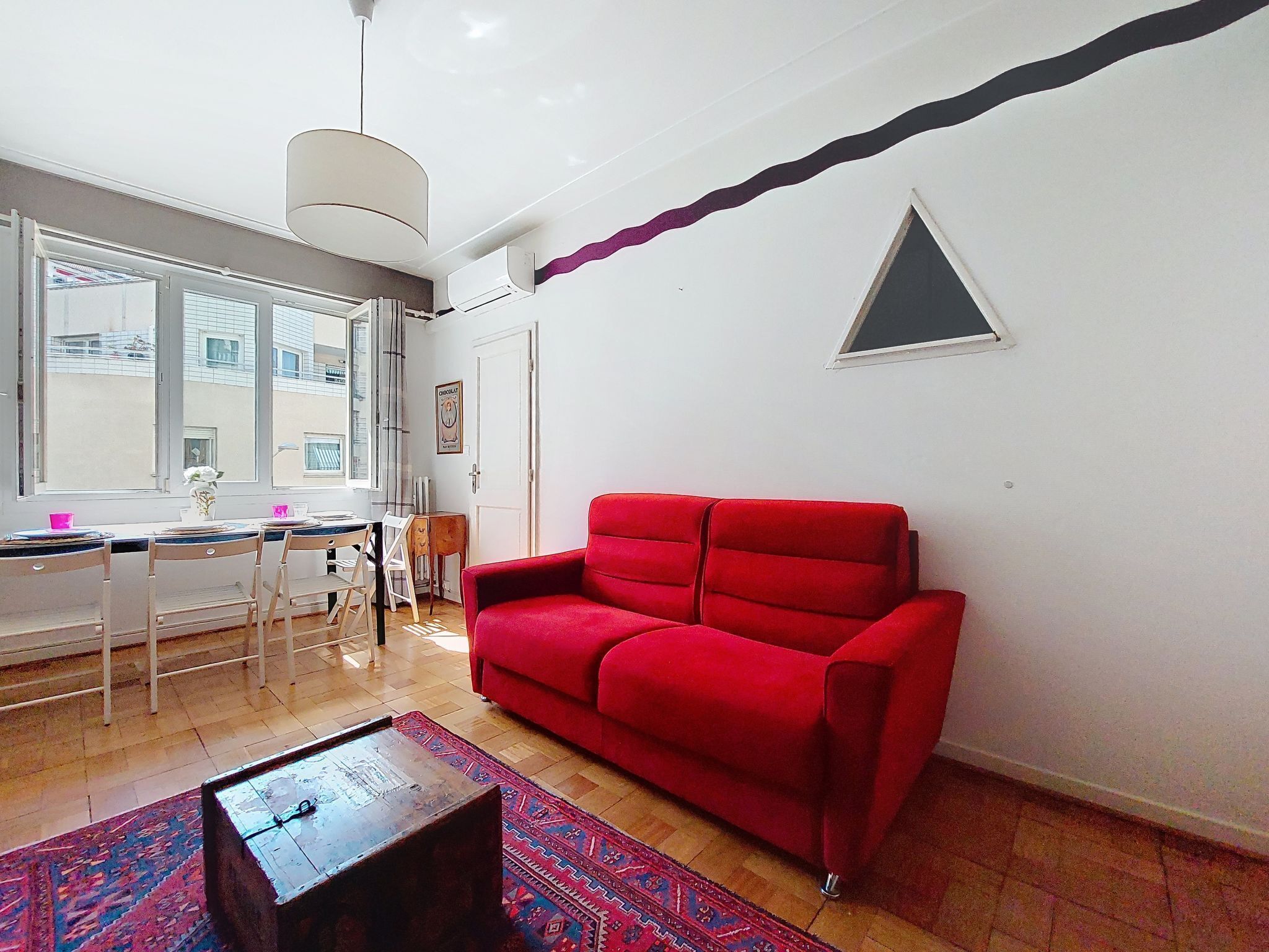 A living room with a red sofa, a dining table with chairs, a coffee table, and a large window with blinds.