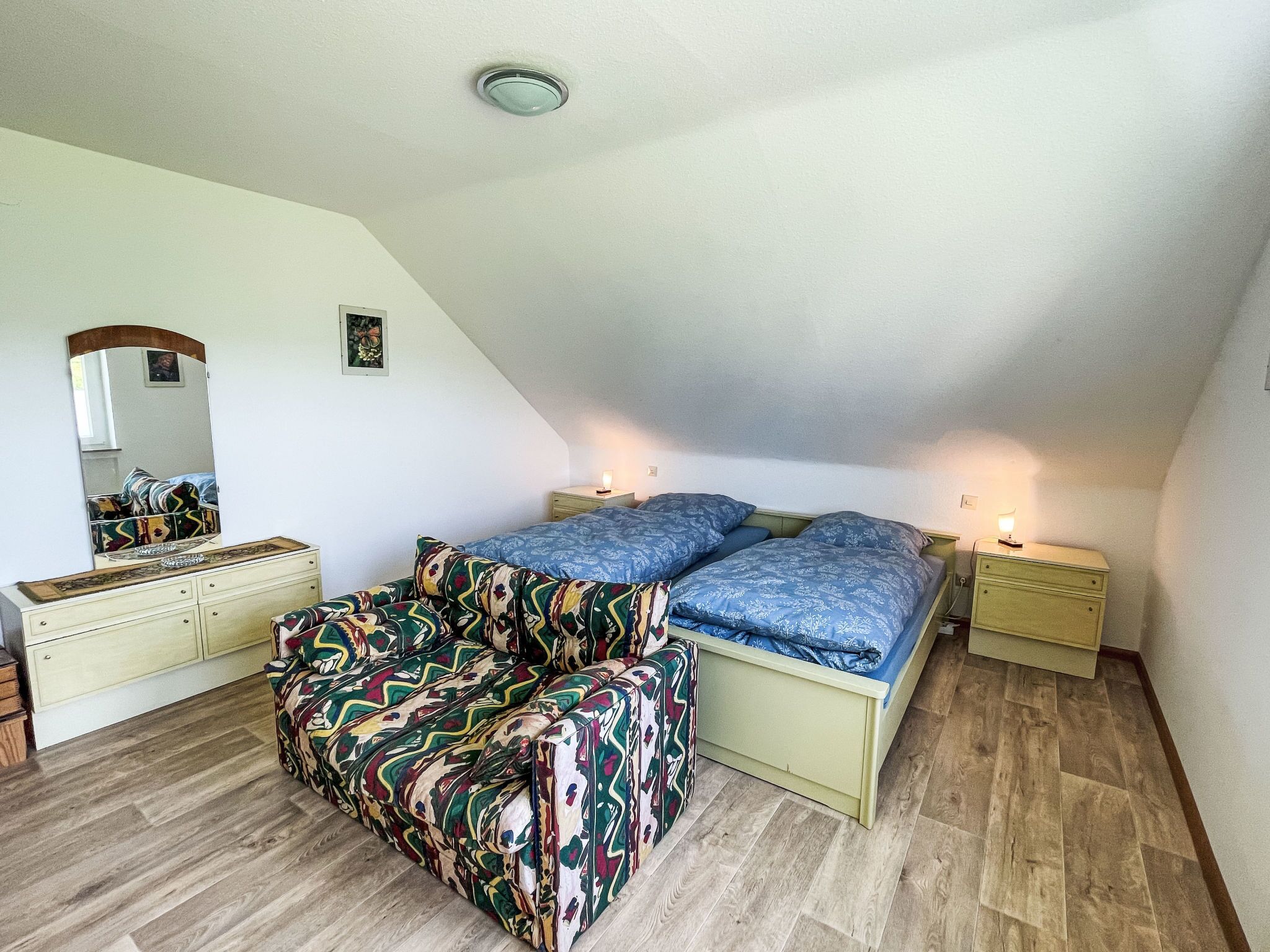 Une chambre avec un plafond en pente, un lit avec une literie bleue, un canapé coloré, une commode et un miroir.