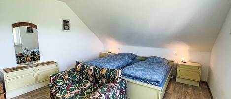 Une chambre avec un plafond en pente, un lit avec une literie bleue, un canapé coloré, une commode et un miroir.