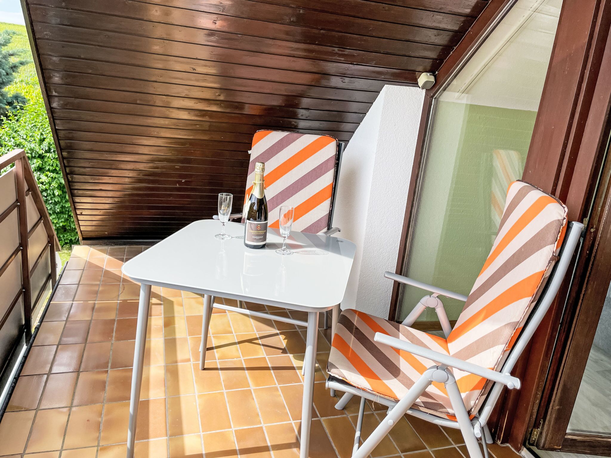 A small patio with a white table and two striped chairs, a bottle of champagne, and a glass on the table.