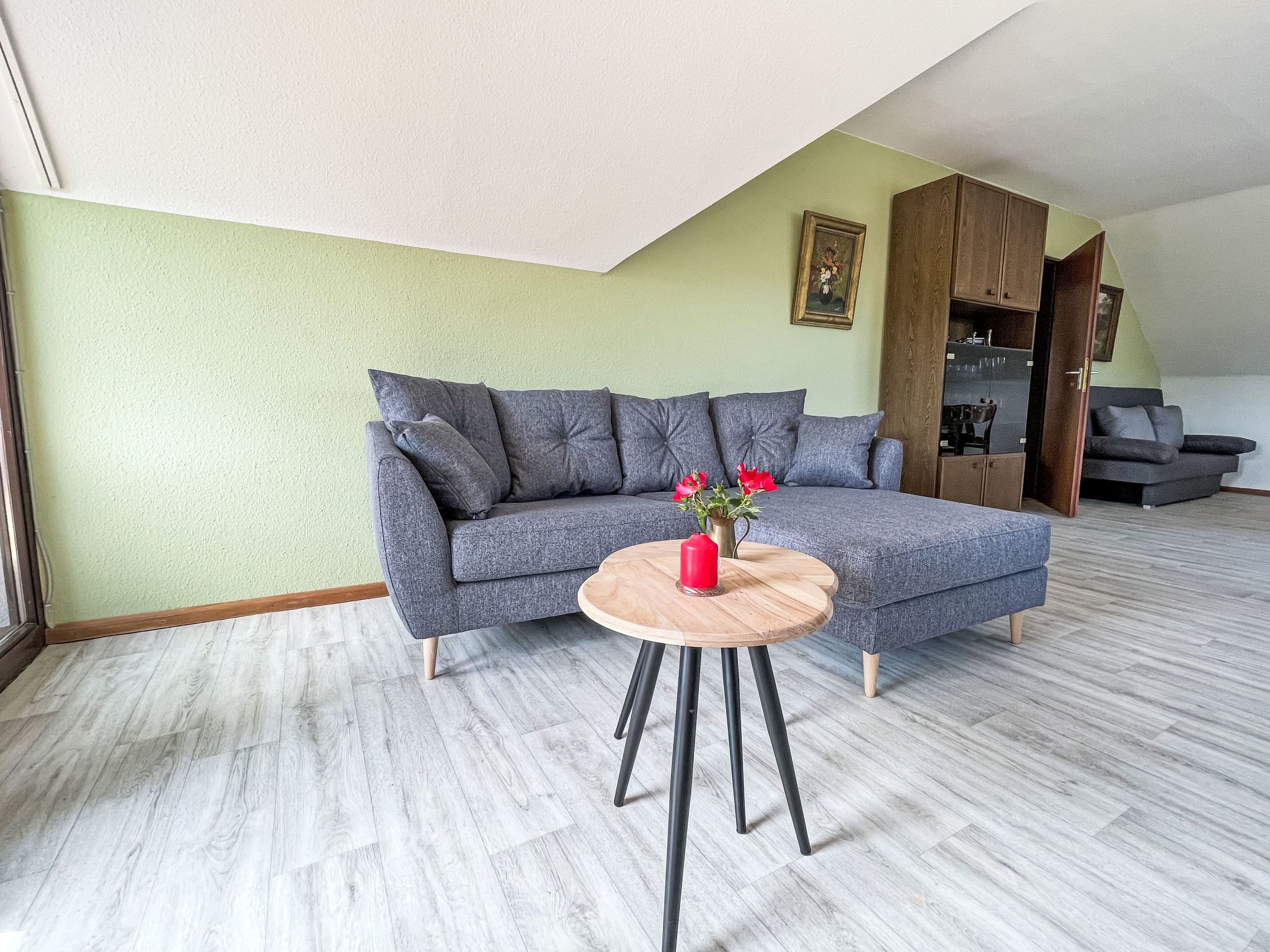 Una sala moderna con un sofá seccional gris, una mesa de centro redonda de madera con una vela roja, y un comedor con un mueble de madera y un sofá gris.