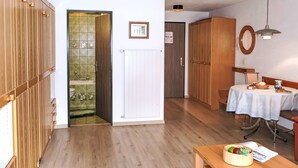 A room with wooden flooring, a white radiator, a wooden door leading to a bathroom, a table with a basket of fruit, and a hanging light fixture. - Pasadena by Interhome (Zermatt)