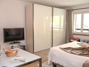 Ein Hotelzimmer mit einem Bett, einem Fernseher, einem Schreibtisch mit einem Brotkorb, einem Weinglas und einem Fenster mit Vorhängen.