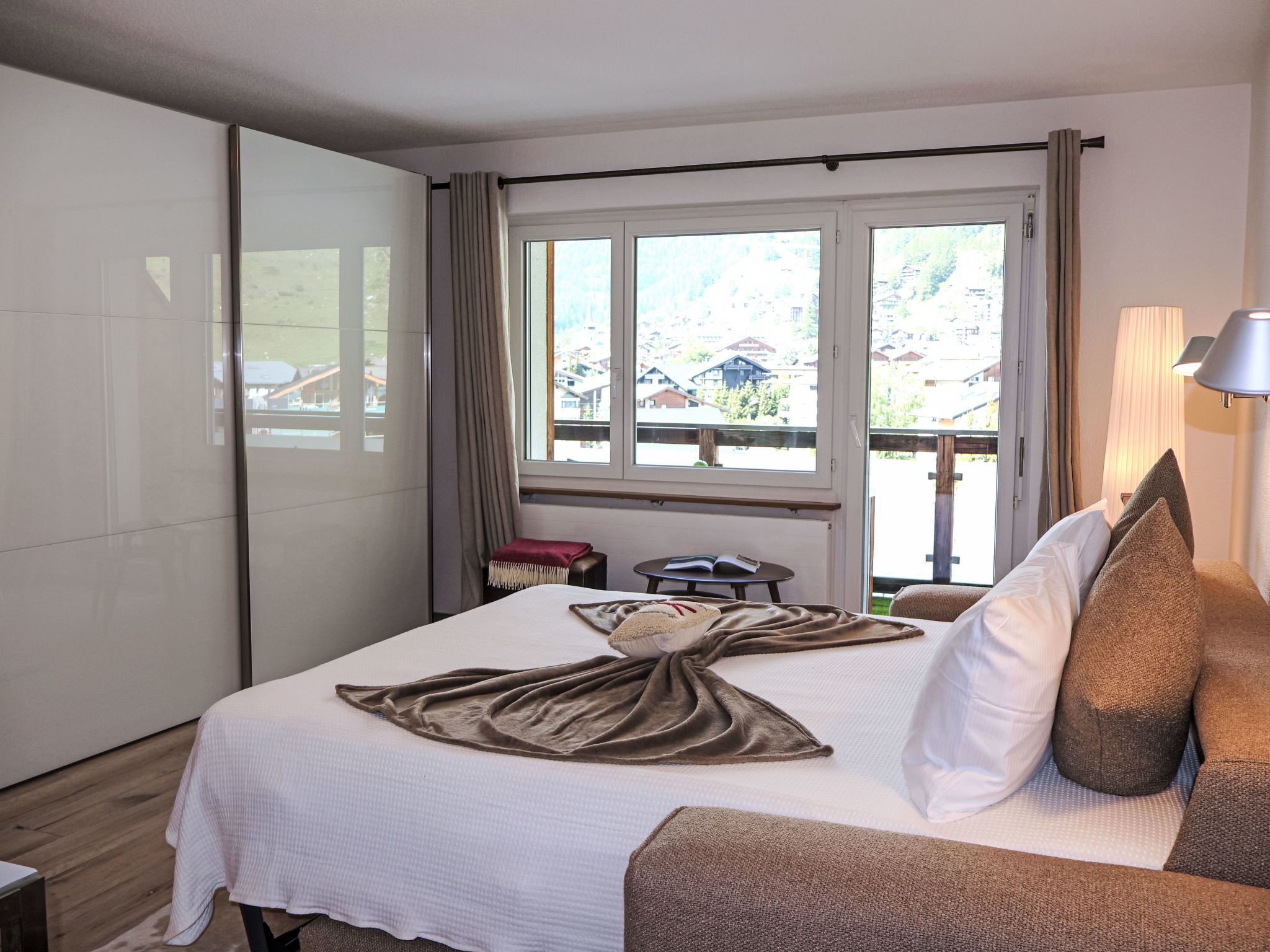 A hotel room with a bed, a small table, a lamp, and a view of the outside.