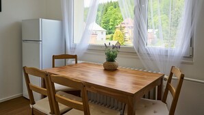 A small kitchen with a white refrigerator, a wooden dining table with four chairs, and a window with sheer curtains. - Forest line by Interhome (Josefuv Dul)