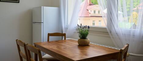 A dining area with a wooden table and chairs, a small potted plant, a white refrigerator, and sheer curtains.