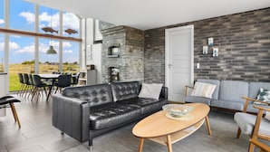 A modern living room with a black leather sofa, a wooden coffee table, and a grey sofa. There is a brick wall and a large window offering a view of the outdoors. - "Ung" - 460m from the sea by Interhome (Otterup)