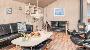 A cozy living room with a wooden ceiling, a black leather sofa, a dining table with fruits and glasses, and a wood-burning stove.