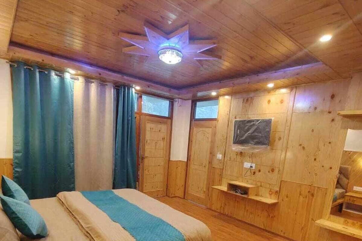 Ένα δωμάτιο με ξύλινο δάπεδο, ένα κρεβάτι, κουρτίνες, μια τηλεόραση τοποθετημένη στον τοίχο και ένα φωτιστικό οροφής.