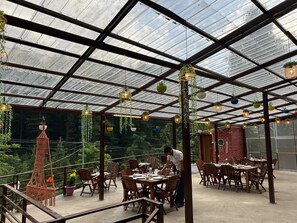 Um restaurante com teto de vidro, mesas de madeira e plantas penduradas.