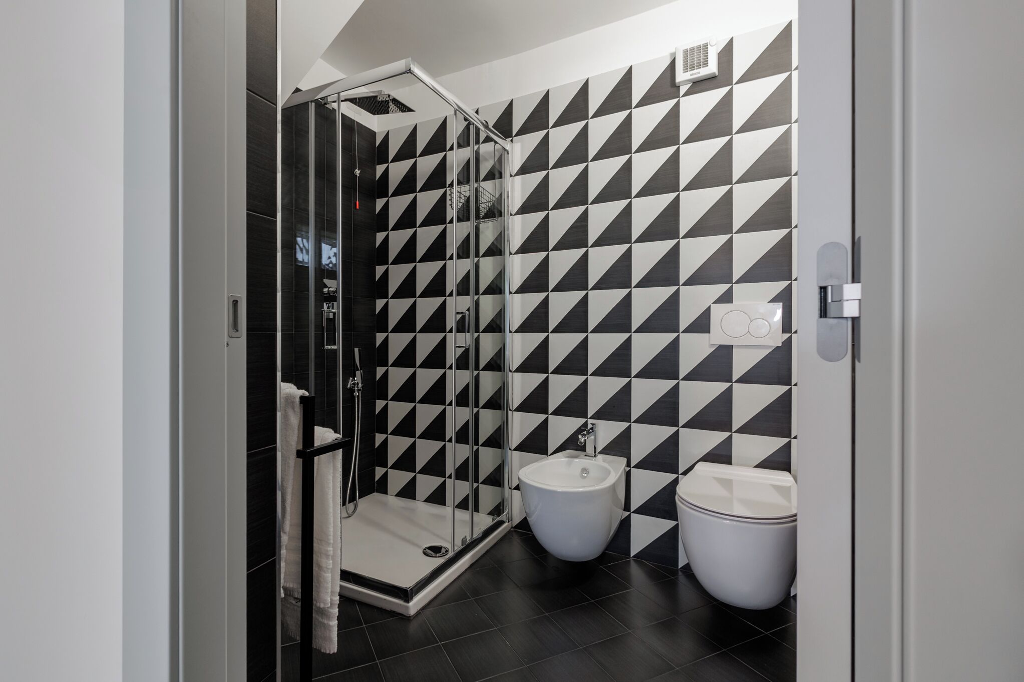 A modern bathroom with a black and white checkered tile wall, a glass-enclosed shower, and two white toilets.