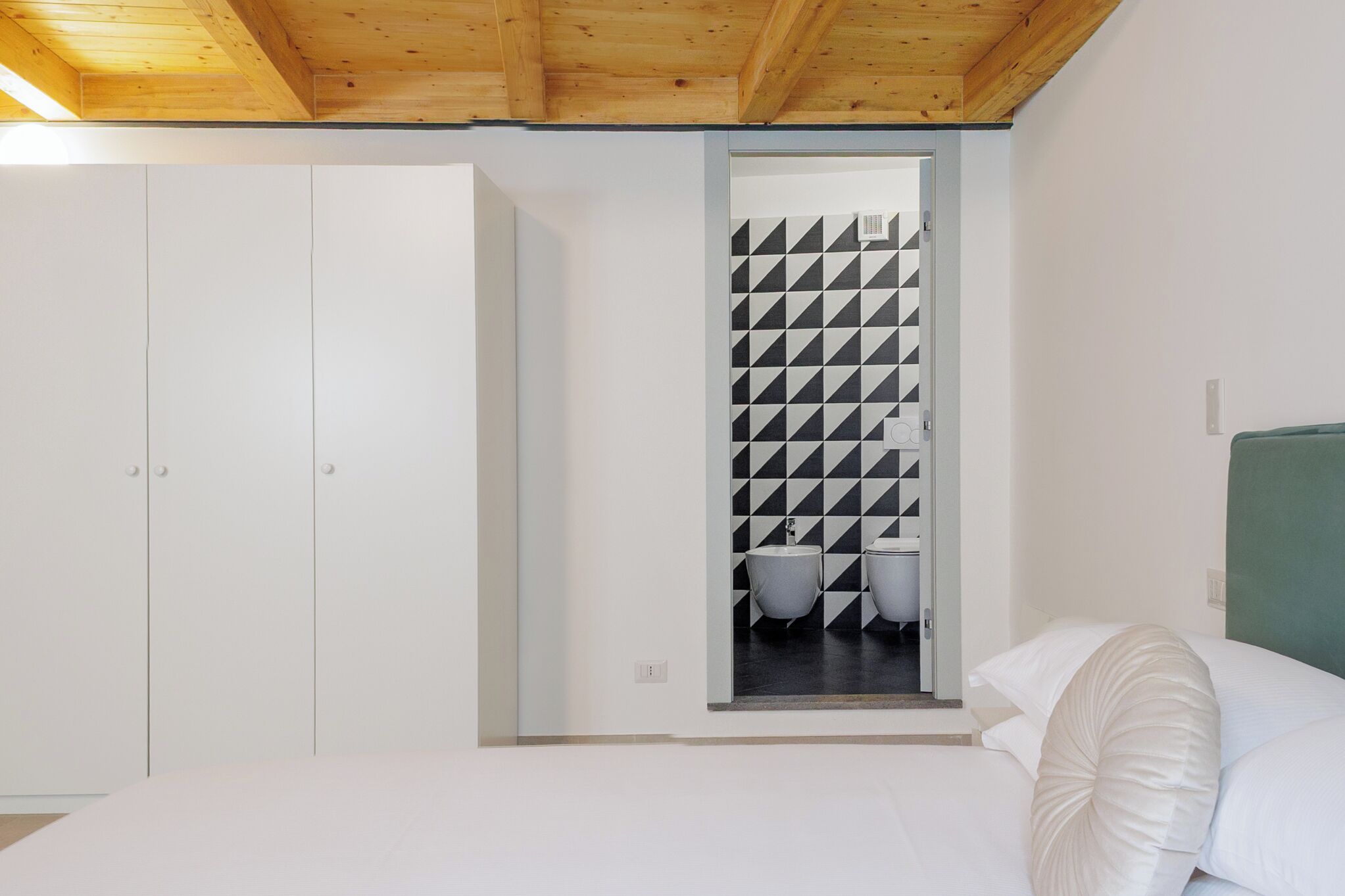 Een slaapkamer met een houten plafond, een witte inloopkast, een bed met witte beddengoed en een badkamer met een zwart-wit geometrisch tegelpaneel.
