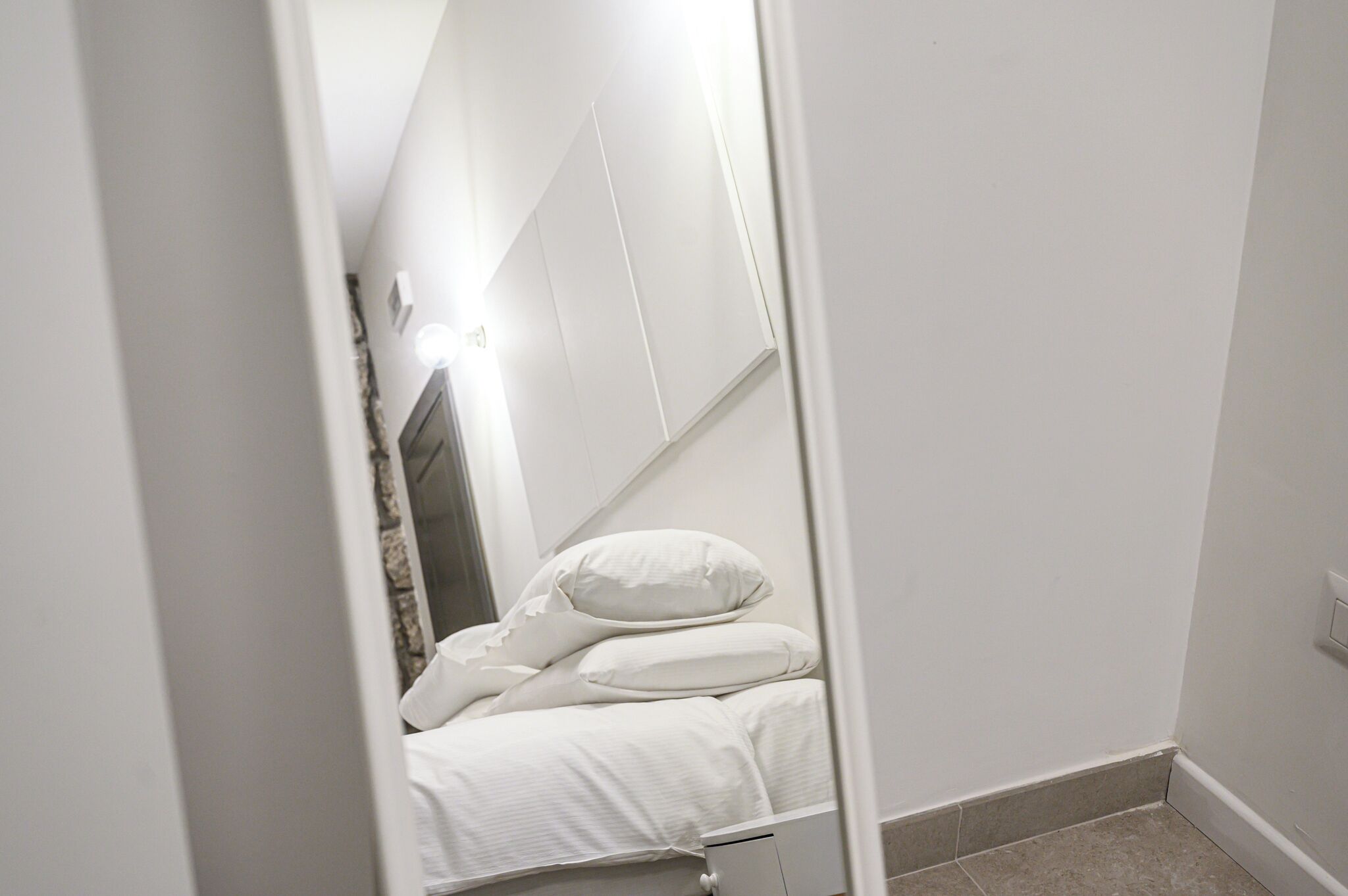 A narrow hallway leading to a bedroom with a bed, pillows, and a nightstand.