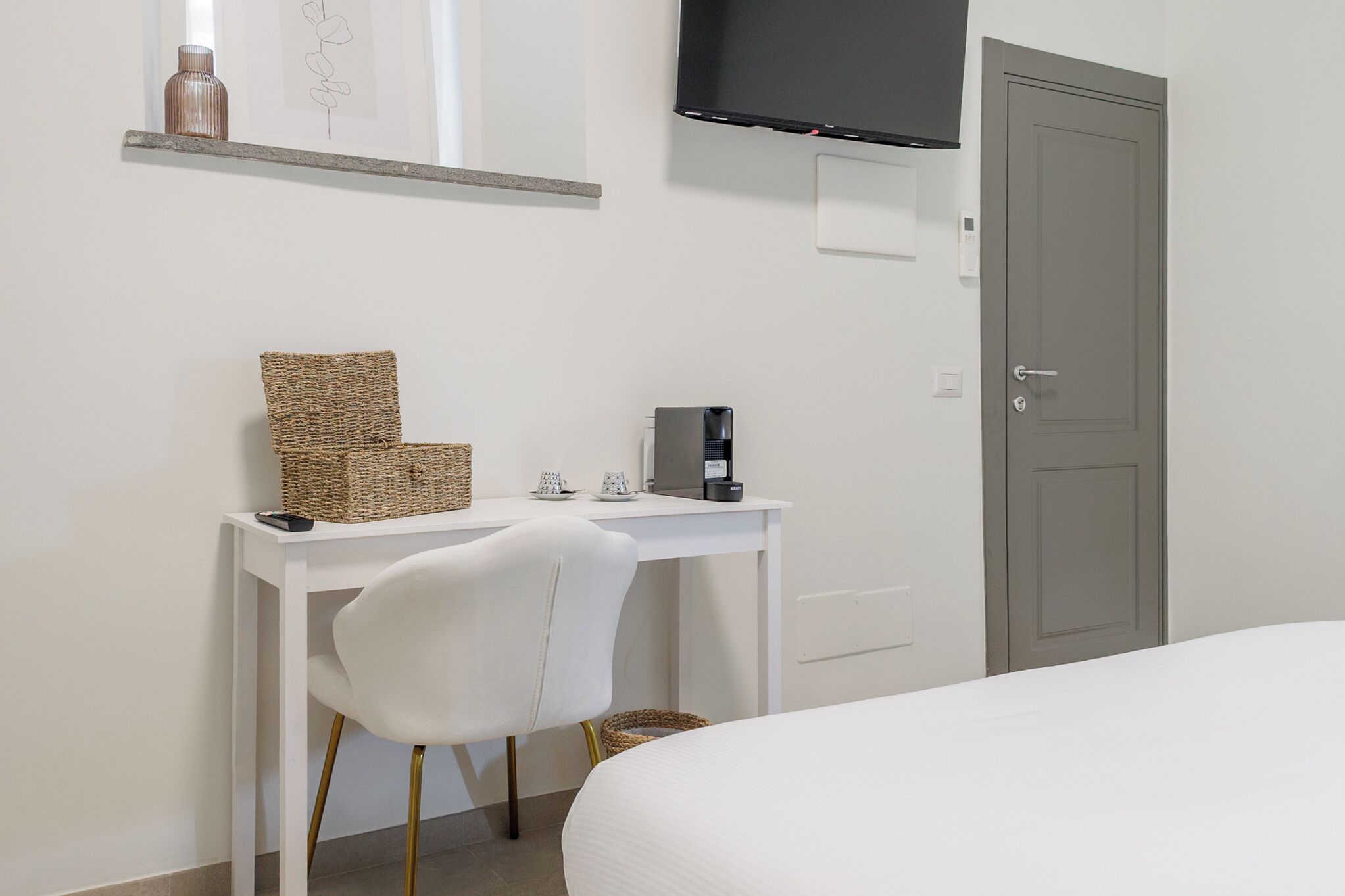A hotel room with a white desk, a white chair, a bed, a television mounted on the wall, and a door.