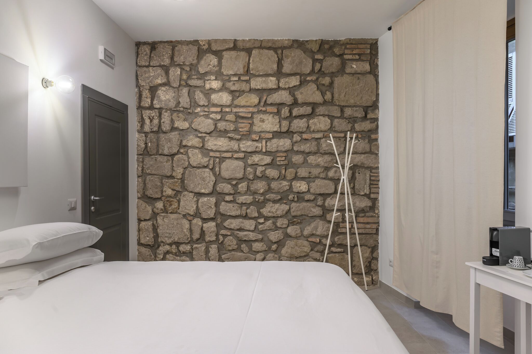 Une chambre avec un mur en pierre, un lit avec une literie blanche, une porte, une table de chevet avec une machine à café et un rideau.