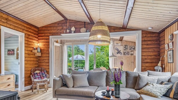 L’intérieur d’un chalet confortable, avec un canapé, un fauteuil en osier, une table ronde et une grande fenêtre donnant sur un paysage verdoyant.