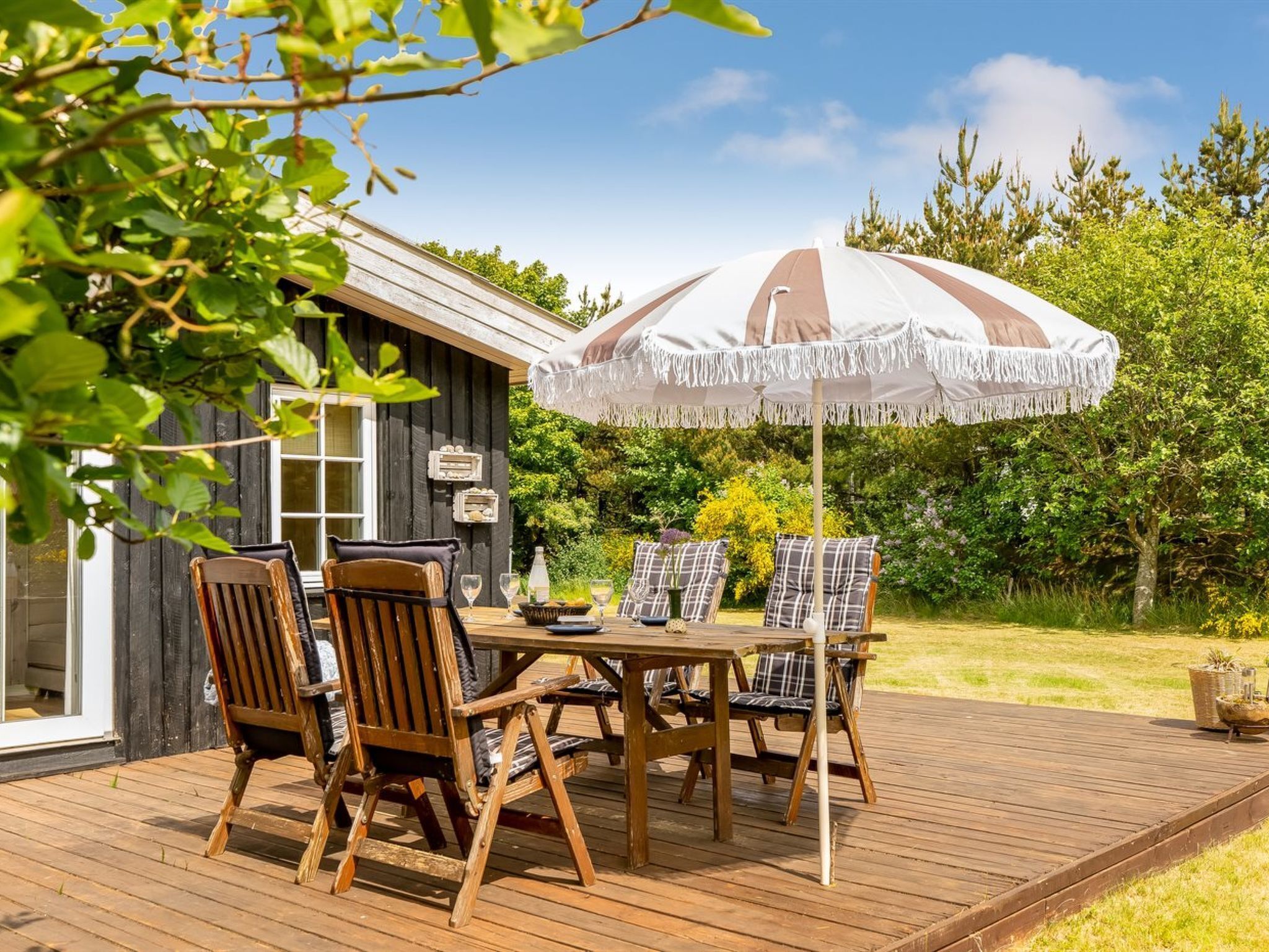 Une terrasse en bois avec une table et des chaises, un parasol et un petit abri de jardin, le tout aménagé dans un cadre verdoyant.